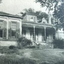 Black and white photo of an old house with a porch and overgrown yard.