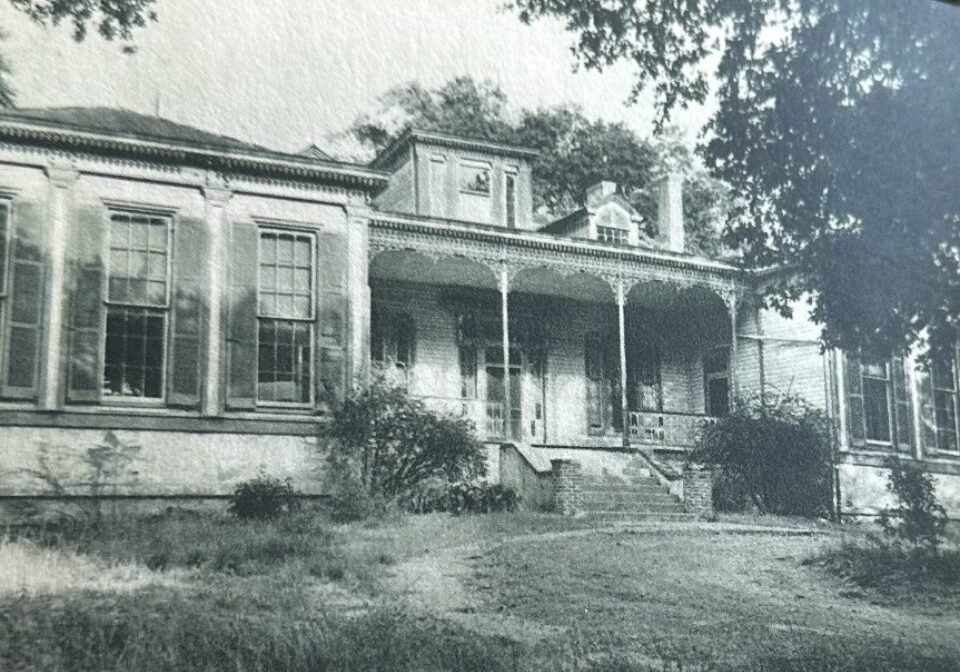Black and white photo of an old house with a porch and overgrown yard.